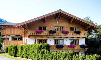 A rustic chalet with wooden siding and a balcony, decorated with colorful flowers. Surrounded by a green hedge and beautiful scenery.