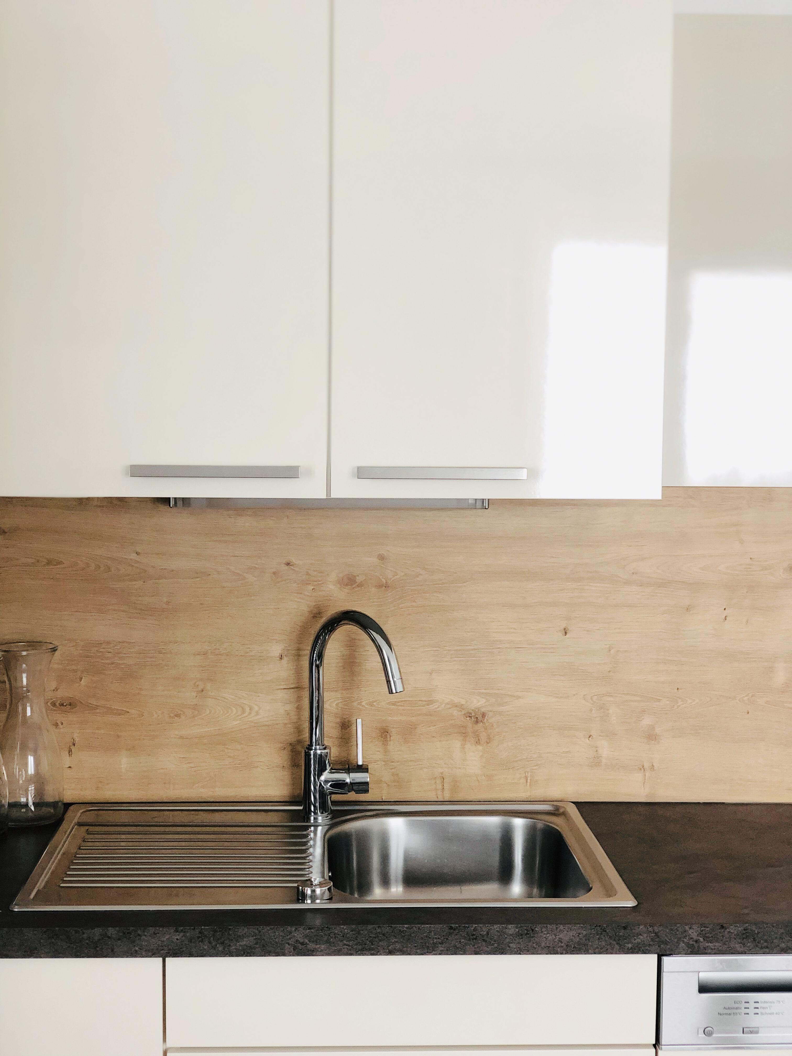 A modern kitchen featuring a stainless steel sink and a simple wood backsplash. The white cabinets create a bright and clean atmosphere.