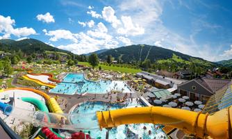 A water park with several swimming pools and slides. In the background, green hills and a blue sky with clouds can be seen.