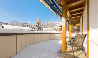 A cozy terrace with a table and chairs, surrounded by snow and a clear blue sky. In the background, snow-covered trees and mountains are visible.