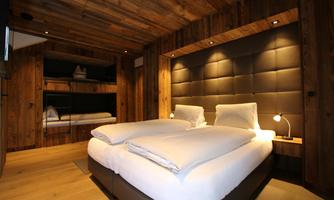 Family-friendly bedroom with a double bed and integrated bunk bed. This spacious chalet room combines stylish design with practical functionality. The large double bed with a padded leather headboard invites relaxation, while the fixed bunk bed made of reclaimed wood, with cozy lighting, particularly delights children. Warm wooden paneling, subtle light sources, and high-quality materials create a cozy atmosphere of well-being – ideal for families with up to two children.