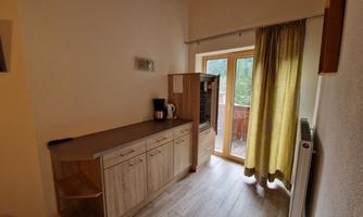 A modern kitchenette with wooden cabinets and a kettle. Large windows with yellow curtains allow plenty of daylight in.
