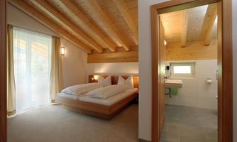 A bright bedroom with a wooden ceiling and two beds. In the background, a modern bathroom is visible.