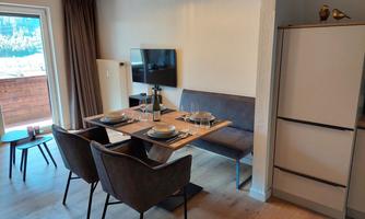 A modern dining room with a table set for dinner. Cozy chairs and a television on the wall are visible.