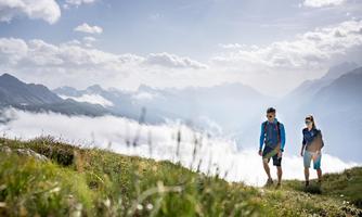 Zwei Wanderer auf einem Berggipfel umgeben von einer malerischen Landschaft. Im Hintergrund sind Wolken und die Berge sichtbar.