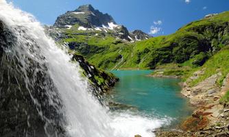 A beautiful waterfall flows into a clear, blue lake. In the background, green mountains rise under a bright blue sky.