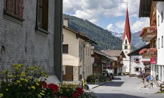 Eine malerische Dorfstraße mit charmanten Häusern und bunten Blumen. Im Hintergrund ragt ein Kirchturm mit rotem Spitzdach hervor, umgeben von Bergen.