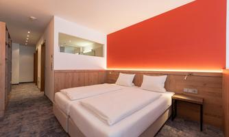 A modern hotel room with a bright, comfortable bed and an elegant red wall. The room is well-lit and friendly designed.