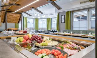A buffet with fresh fruit, vegetables, and cold cuts. The room has an inviting atmosphere with green curtains and bright light.