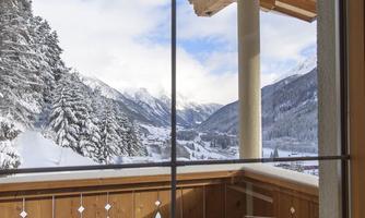 A winter balcony with a wooden railing and a view of snow-covered mountains. The landscape is peaceful and framed by tall fir trees.