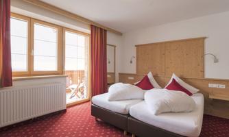 Bright hotel room with two single beds and large windows. Red curtains and wooden furniture create a cozy atmosphere.