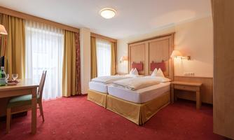 A cozy hotel room with two single beds and a desk. The room has large windows with curtains and a warm color tone.