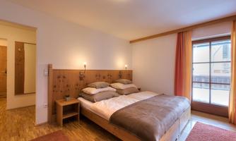 A bright bedroom with a double bed and two nightstands. Large windows with curtains provide plenty of natural light.