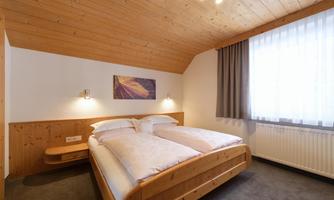 A cozy bedroom with a wooden double bed. The walls are bright, and there is plenty of natural light coming through the window.