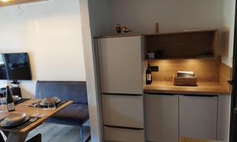 A modern kitchen with a refrigerator and wooden shelves. In the background, a dining table with dishes and a television is visible.