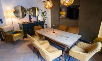 A modern living room with a large wooden table and elegant yellow chairs. Mirrors hang on the walls and there are decorative plants.