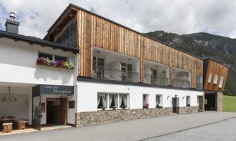 A modern hotel building with wood cladding and several balconies. In the background, green mountains can be seen.