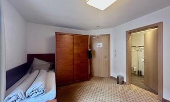 A simple room with a single bed and a large wardrobe. On the right side, there is a bathroom with modern amenities.