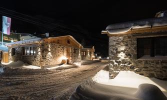 A winter landscape with snow-covered buildings at night. Soft light illuminates the path through the snowy surroundings.