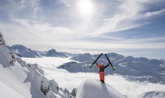 A skier stands triumphantly on a summit in the mountains. Surrounded by snow-covered landscapes and a bright blue sky.