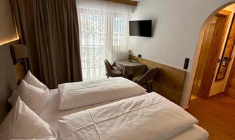 A modern hotel room with two beds and large windows. In the background, a table and a television can be seen.