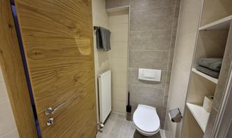 A modern bathroom with white tiles and a wooden interior door. It includes a toilet, towels, and a shelf with gray towels.