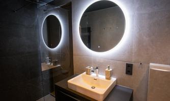 A modern bathroom with an elegant sink and a backlit round mirror. The walls are in neutral gray tones.