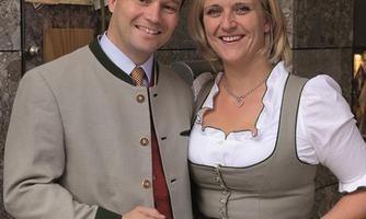 A couple in traditional Bavarian attire poses smiling in front of a stone background. The man is wearing a jacket with buttons and the woman is wearing a dirndl with a striped apron.