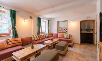A cozy living space with comfortable seating and wooden shelves. Bright windows and decorative curtains create an inviting atmosphere.