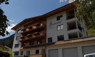 A modern building with several floors and balconies. The façade is bright, surrounded by green nature and a blue sky.