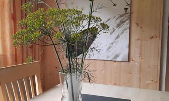 A vase with fresh dill branches is on a table.  
In the background, there is a picture of a skier on a wall.