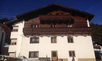 A traditional alpine house with wooden cladding and balconies. It stands in a snowy environment under a clear blue sky.