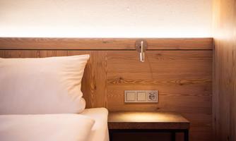 A cozy bed with white pillows and a wooden nightstand. Light sources and power outlets are visible on the wall.