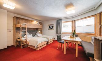 A cozy room with two beds and a bunk bed construction. There is a table and chairs as well as large windows that let in plenty of light.
