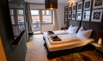 A cozy hotel room with two beds and modern furnishings. Artworks hang on the wall, and the windows provide access to a balcony.