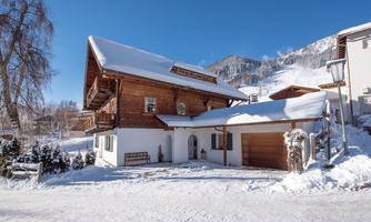 A charming wooden house in winter with a snow-covered roof. In the background, snowy mountains and a blue sky can be seen.