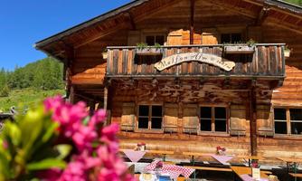 Eine rustikale Berghütte mit einem Holzbalkon und stilvollem Schild. Draußen sind Tische mit karierten Tischtüchern und Blumen dekoriert.