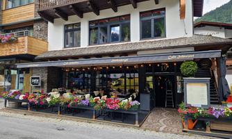 An inviting café with blooming plants on the outdoor terrace. The building has large windows and is surrounded by beautiful mountain landscapes.