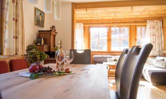 A cozy dining area with a large wooden table, decorated with fruit and glasses. In the background, a bright living space with windows and a sofa can be seen.