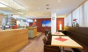 A modern café with wooden decor and cozy seating. The atmosphere is inviting and bright, featuring a decorative wall and plants.