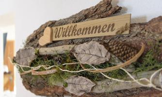 A wooden sign with the inscription "Welcome" and decorative elements such as leaves and pine cones. The background consists of natural tree bark.