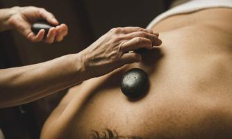 A person is receiving a massage with hot stones. The hands of a therapist are placing a smooth, black stone on the client's back.