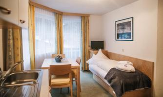 A small, cozy room with a single bed and a table. The windows are adorned with curtains and there is a small kitchenette.