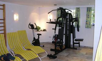 A fitness room with two reclining chairs and a training machine. In the background, there is a stationary bike and bright windows with curtains.