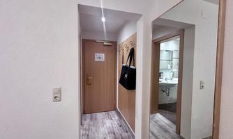 A bright hallway with a mirror and a door to a bathroom. Coat hooks hang on the wall and a bag is visible.