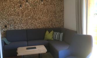 A cozy living room with a blue sofa and a wooden table. The walls are decorated with wooden logs and plenty of natural light streams through the window.