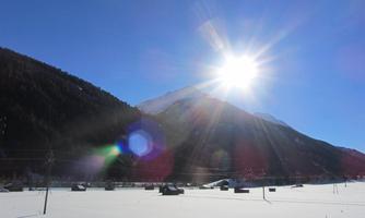 A snowy landscape with mountains in the background. The sun shines brightly over the scene.