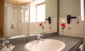 A modern bathroom with a sink and a shower. On the table, there are flowers in a vase, and there is plenty of natural light.
