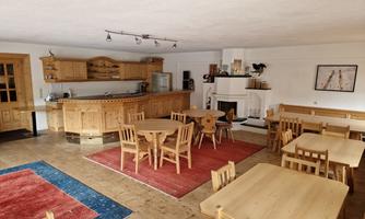A cozy room with wooden chairs and tables. In the background, there is a kitchen and a fireplace.