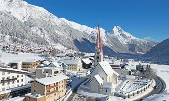 A picturesque winter landscape with snow-covered mountains and a small village. The church with the red roof is the focal point of the scene.